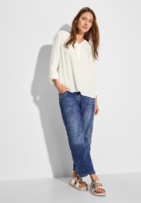 Blusa di colore crema, dal taglio morbido, con scollatura a bottoni, indossata con jeans in denim blu e sandali argentati a doppia fibbia.