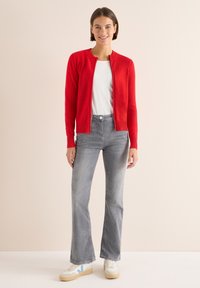 Cardigan rosso a maglia con bordi a coste, indossato su una camicia bianca, abbinato a jeans grigi svasati e sneakers bianche con accenti blu.