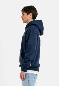 Felpa blu navy con tasca a marsupio, polsini e fondo a coste, e logo ricamato bianco sul davanti e sulla manica. Tessuto morbido.