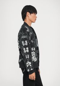 Giubbotto bomber nero con stampe floreali e farfalle, dotato di chiusura a zip frontale, polsini a costine e una texture di tessuto liscia. Vestibilità slim.