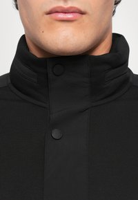 Veste noire à col haut en tissu lisse, dotée d'un design structuré, de deux boutons-pression noirs et de détails de couture visibles.