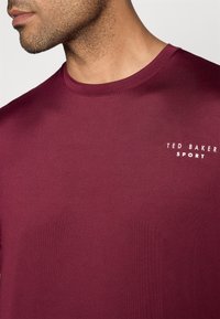 Burgundská tričko s krátkym rukávom vyrobené z bavlny. Má okrúhly výstrih a biele logo "TED BAKER SPORT" na ľavej strane.