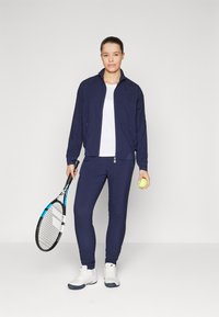 Navy athletic jacket and pants, white shirt, holding a tennis racket and ball. Shoes are white with dark soles. Smooth fabric, fitted design.