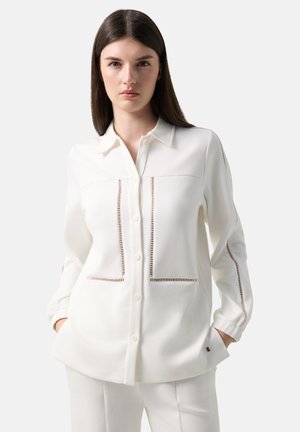 Frau mit langen dunklen Haaren trägt ein weißes Hemd mit Knöpfen und dekorativen Ausschnitten vorne und an den Ärmeln, Hände in den Taschen.