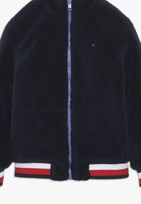 Granatowa polarowa kurtka na suwak z kapturem, z niebieskim zamkiem błyskawicznym, prążkowanym dołem z czerwono-białymi paskami oraz małym logo na piersi.