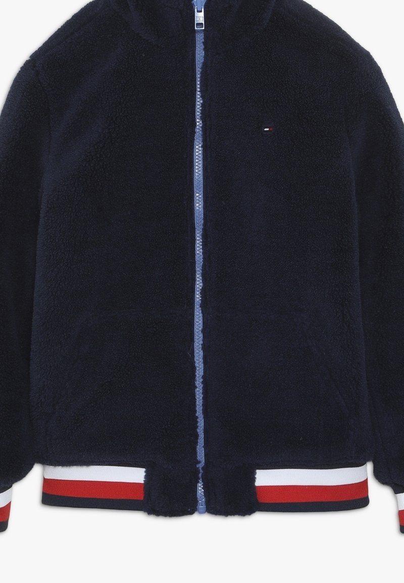 Granatowa polarowa kurtka na suwak z kapturem, z niebieskim zamkiem błyskawicznym, prążkowanym dołem z czerwono-białymi paskami oraz małym logo na piersi.