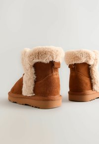 Brune ruskinds støvler med lyse kunstpelsfoder og tan gummisåler. Designet har en rund tå og en lav ankelhøjde.