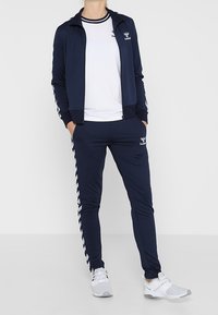 Marineblaue Trainingsanzug mit Reißverschlussjacke und passenden Hosen. Verfügt über weiße Streifenakzente an Ärmeln und Hosen. Kombiniert mit grauen Sportschuhen.