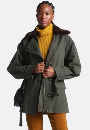 Femme aux cheveux tressés porte un manteau vert olive avec un col en fourrure sur un pull à col roulé côtelé moutarde et un pantalon moutarde, tenant les revers du manteau.
