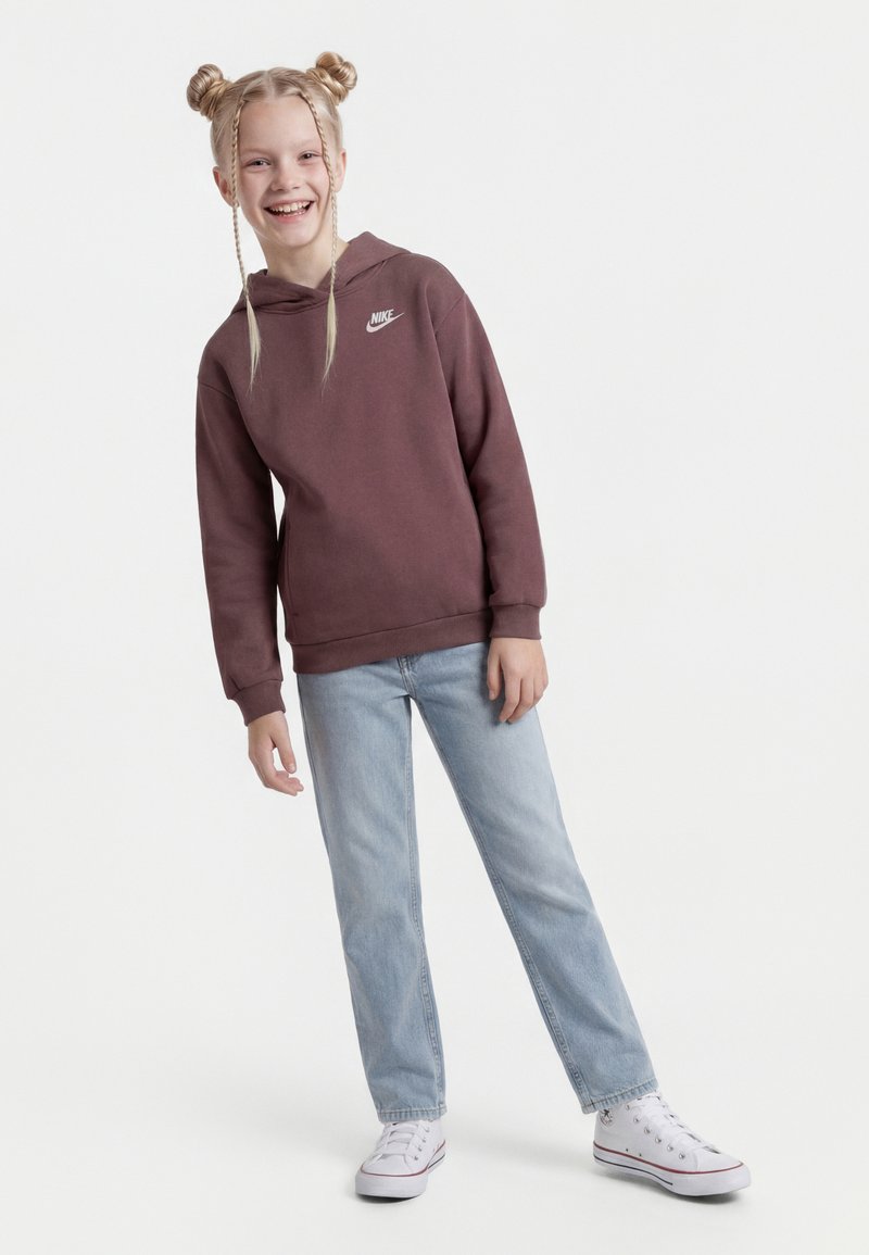 Χαμογελαστή κοπέλα με δύο πλεξούδες σε σχήμα κότσου, ντυμένη με μπορντό φούτερ Nike, ανοιχτό μπλε τζιν και λευκά αθλητικά παπούτσια, που στέκεται μπροστά σε λευκό φόντο.