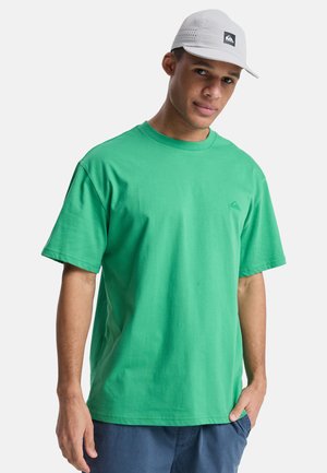 Młody mężczyzna w jasnoszarej czapce, zielonej koszulce z małym logo na piersi oraz ciemnoniebieskich szortach, stojący z jedną ręką w kieszeni.