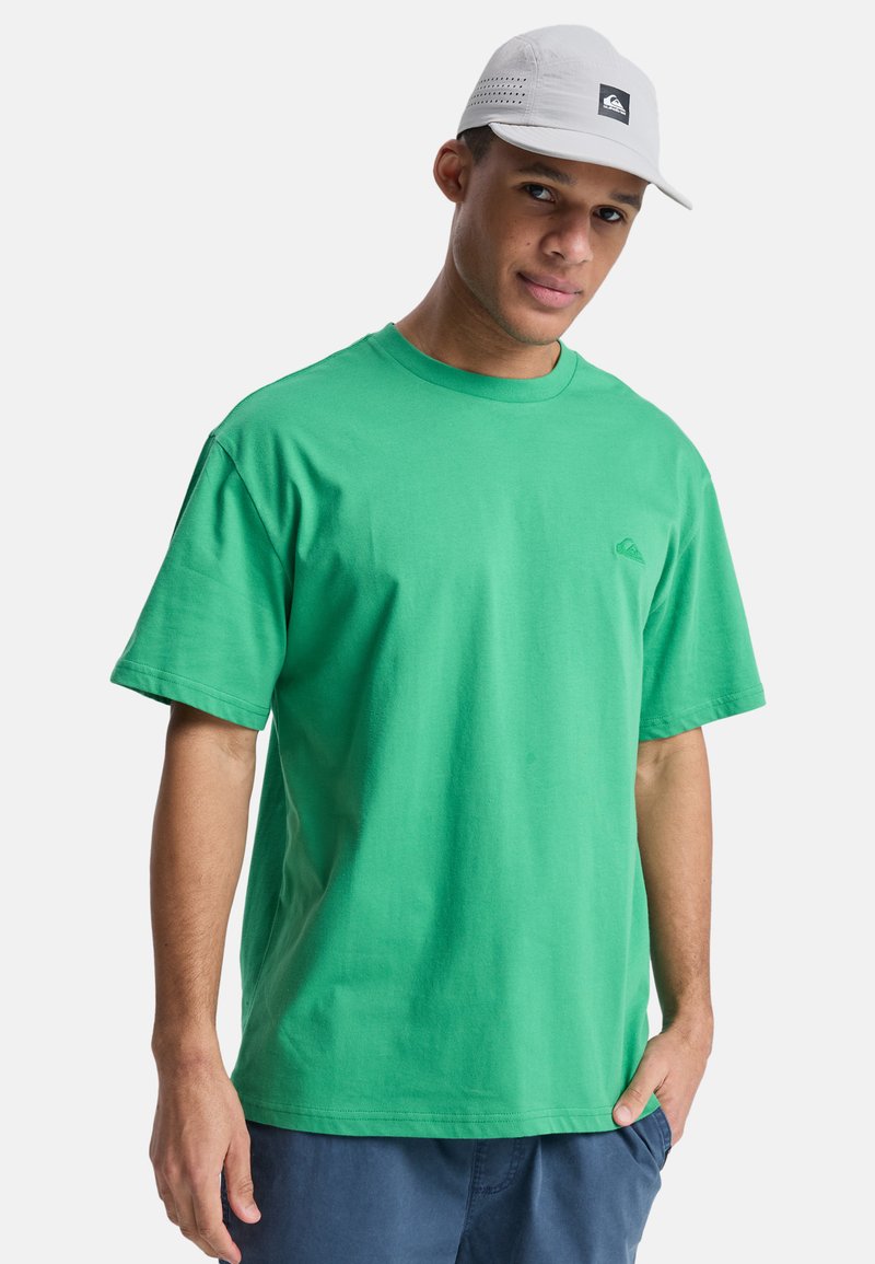 Mladý muž s svetlosivou čiapkou, zeleným tričkom s malým logom na hrudi a tmavomodrými šortkami, stojaci s jednou rukou vo vrecku.