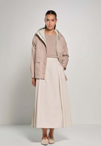 Lichte, beige jas met een capuchon, gedragen met een beige top en een lange, vloeiende witte rok. Soepele textuur, eenvoudig ontwerp.