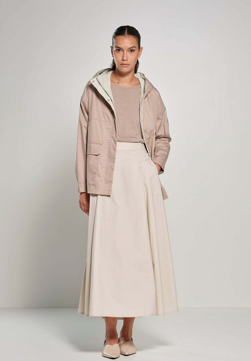 Lichte, beige jas met een capuchon, gedragen met een beige top en een lange, vloeiende witte rok. Soepele textuur, eenvoudig ontwerp.