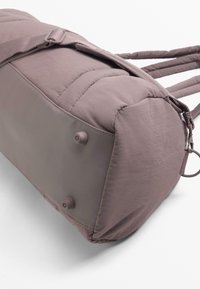 Sac de voyage matelassé mauve avec une bandoulière réglable, base plate avec deux pieds de protection sur fond blanc.