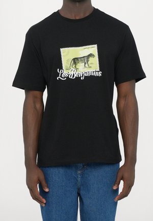 Schwarzes T-Shirt mit einem gelben Briefmarkenmotiv eines anatolischen Leoparden über dem weißen Schriftzug "Les Benjamins", getragen von einer Person in blauen Jeans.