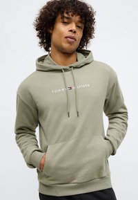 Mladý muž s kučeravými vlasmi, oblečený v svetlozelenej mikine Tommy Hilfiger, s rukami v prednom vrecku, biely pozadie.