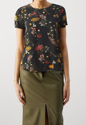Vrouw draagt een zwart kortärmelig blouse met een meerkleurige bloemenprint en een olijfgroene rok met een voorsplit.