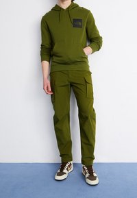 Zielona bluza z kapturem z przednią kieszenią i logo, zestawiona z dopasowanymi spodniami bojówkami z bocznymi kieszeniami i luźnym krojem, noszona z brązowo-białymi trampkami.