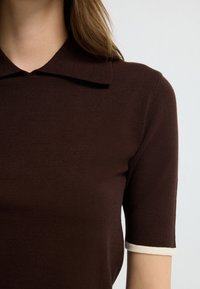 Brun strikket polo-shirt med krave og korte ærmer. Ærmerne har en cremefarvet kant. Glat tekstur, tætsiddende design.