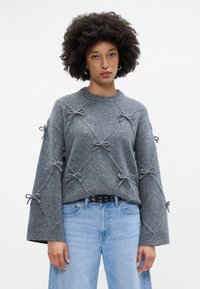 Pilkas megztinis su varpo formos rankovėmis, papuoštas dekoratyvinėmis juostelėmis visame paviršiuje ir apvaliu iškirpimu. Dėvimas su šviesiai mėlynomis džinsomis.