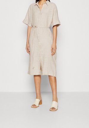 Femme portant une robe-chemise beige boutonnée avec une fine ceinture et des sandales crème à talon carré et bout ouvert sur fond blanc.