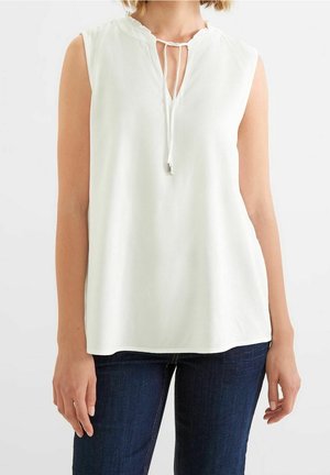 Sleeveless white blouse with a tie at the neckline worn by a person with dark blue jeans against a plain background.