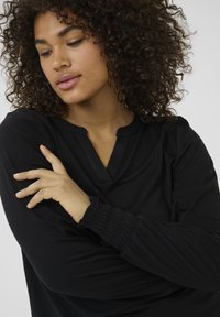 Haut noir à manches longues avec un col en V et des détails froncés aux poignets. Tissu lisse avec une coupe décontractée et sans motifs visibles.