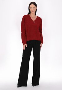Pulover tricotat roșu, pufos, cu decolteu în V, asociat cu pantaloni negri, largi. Modelul stă pe un fond alb, purtând bijuterii simple.