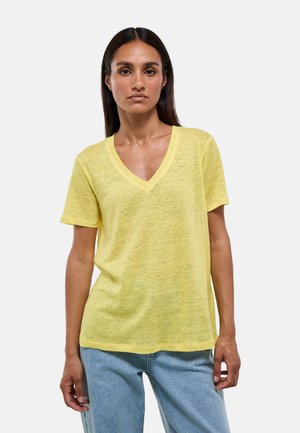 Femme aux longs cheveux foncés portant un t-shirt jaune à manches courtes et col en V, et un jean bleu clair, debout devant un fond uni.