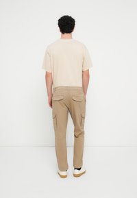 Μπεζ βαμβακερό t-shirt και ταμπά παντελόνι cargo με πολλές τσέπες, άνετη εφαρμογή και ελαστικά μανσέτα. Άσπρα αθλητικά παπούτσια με λαστιχένιες σόλες.