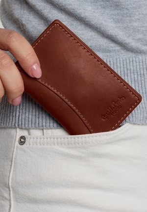 Brown leather wallet with stitching and embossed logo, half-visible in a pocket, paired with a light gray ribbed sweater and white pants.