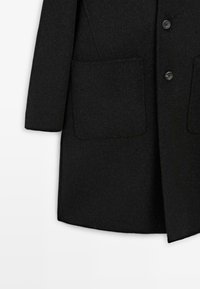 Cappotto di lana nero con un design classico, bottoni frontali e due grandi tasche a toppa con una texture liscia. Silhouette su misura, lunga fino al ginocchio.