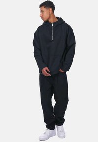 Schwarze Zip-Hoodie mit Kängurutasche, aus weichem Stoff. Kombiniert mit lockeren schwarzen Hosen und weißen Sneakern.