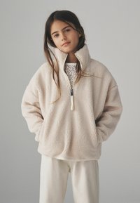 Ljusbeige fleece-pullover med hög krage och halvdragkedja. Mjuk textur, oversized passform och kängurufickor. Lager på en mönstrad undertröja.