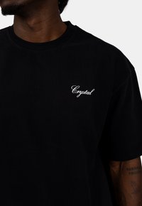 Czarna bawełniana koszulka z krótkim rękawem i okrągłym dekoltem, z białym haftowanym napisem "Crystal" po lewej stronie klatki piersiowej.