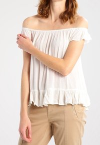 Bílý off-shoulder top s volánkovým lemováním, vyrobený z lehkého materiálu. Modelka má na sobě světle béžové kalhoty, které ukazují uvolněný střih a ležérní styl.