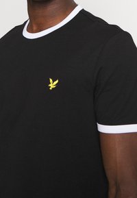 Czarny t-shirt z białym żebrowanym dekoltem i mankietami, z małym haftowanym żółtym logo orła na piersi. Bawełnowy materiał, luźny krój.