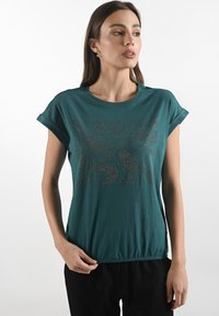 Žena v tyrkysovém tričku s krátkým rukávem a dekorativním vzorem na přední straně, postávající s rukou na boku na bílém pozadí.