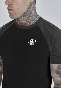 Schwarzes T-Shirt mit kurzen Ärmeln und Raglanärmeln sowie einem kleinen weißen Logo auf der Brust. Der Stoff wirkt glatt und hat eine taillierte Passform.