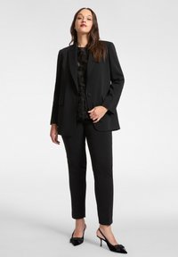 Schwarzes Anzugset mit einem maßgeschneiderten Blazer mit Reverskragen, kombiniert mit eng anliegenden Hosen und schwarzen Slingback-Pumps. Darunter ein strukturiertes Top.