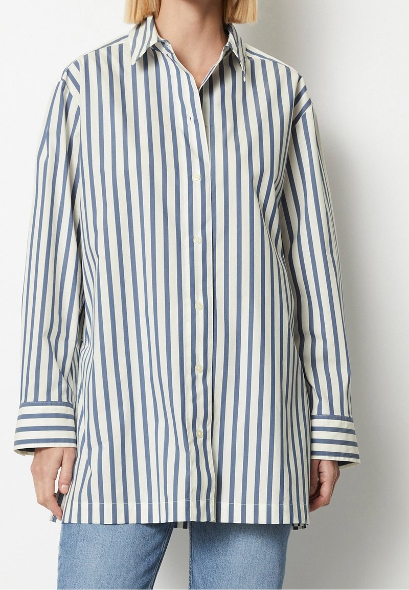 Chemise à manches longues avec des rayures verticales bleu marine et blanches, fermeture boutonnée devant et larges poignets, portée avec un jean bleu clair.