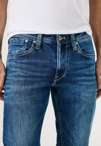 Blå denimjeans med medelhög midja, klassisk femfickdesign, subtila blekningar och gula sömnadsdetaljer vid midjebandet.