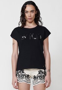 Camiseta de algodón negra con un logo texturizado de "WILO" en lentejuelas metálicas. Atuendo combinado con pantalones cortos crema que presentan bordados florales en negro.