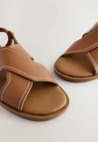 Sandali in pelle marrone con fasce incrociate e cuciture bianche, punte aperte e suola piatta, progettati per comfort e traspirabilità.