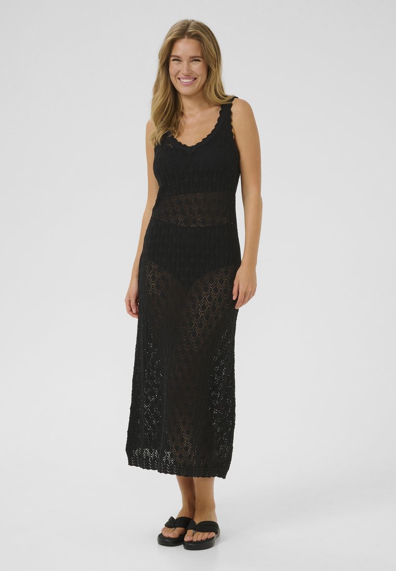 Schwarzes Maxi-Kleid aus Häkelmaterial mit ärmellosem Design, gewelltem Ausschnitt und diamantförmigem Muster. Getragen mit schwarzen Sandalen.