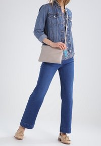 Woman wearing denim jacket and jeans, holding a small beige crossbody bag, standing with one leg slightly forward against a plain background.