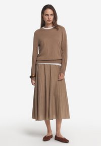 Ljusbrun stickad tröja lagd över en vit skjorta, parat med en plisserad midikjol med guldtrådar och bruna lägenheter. Enkla guldörhängen.