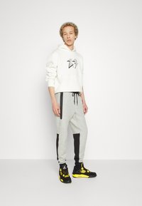 Witte hoodie met een zwart grafisch logo, gecombineerd met grijze sweatpants met zwarte accenten aan de zijkant en met ritszakken. Zwart en gele sneakers.