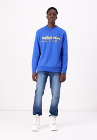 Modrá mikina s logem "Red Bull Racing" v žluté barvě, kulatý výstřih, žebrované manžety, v kombinaci s světle modrými džíny a bílými teniskami.